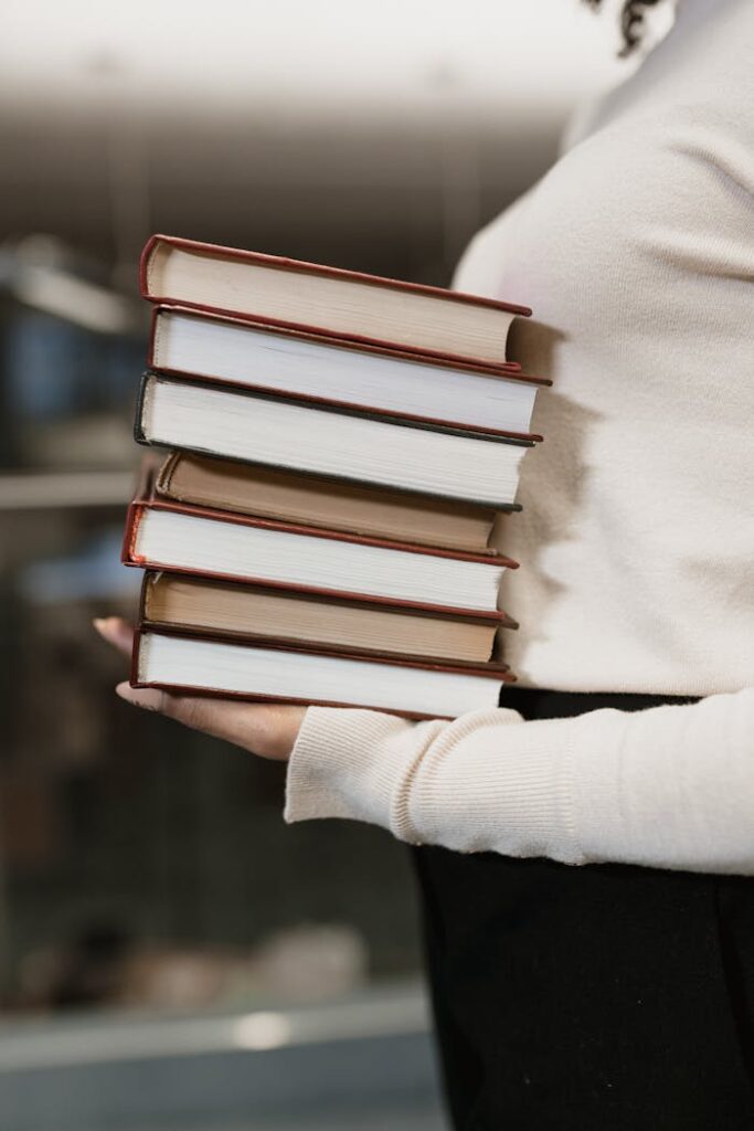 A woman holds a stack of hardcover books, wearing a sweater indoors, captured in a vertical shot.